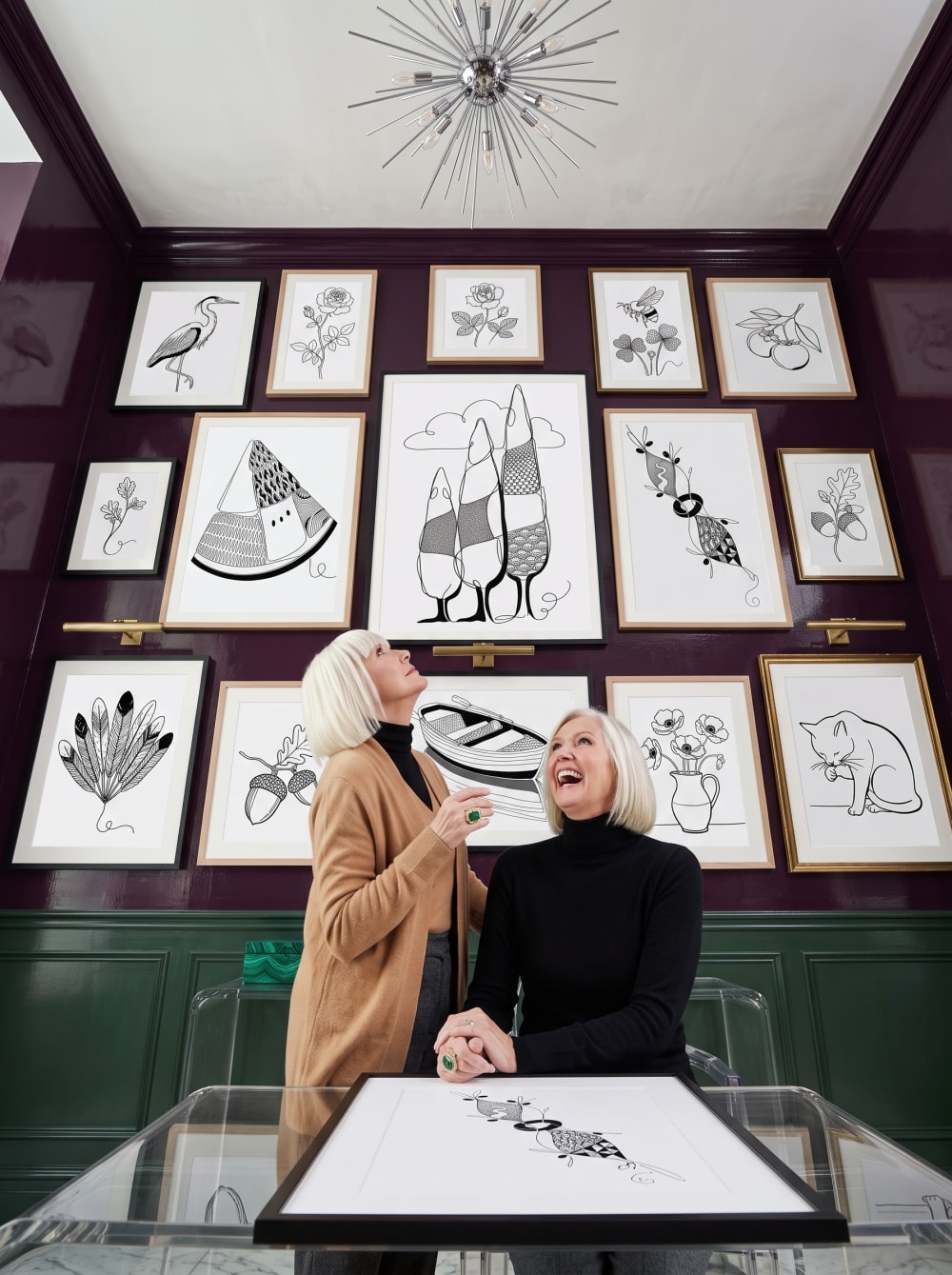 A plum gallery room hung floor-to-ceiling with one-line ink drawings — heron, roses, acorns, a rowing boat, a cat — as two women look up at the wall, laughing