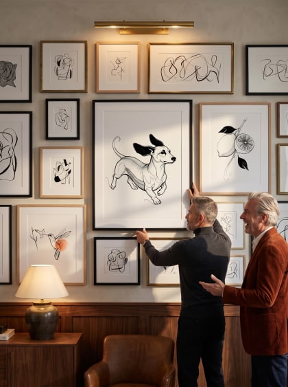 A walnut study salon wall — the dachshund at center, two men mid-laugh as one straightens the frame