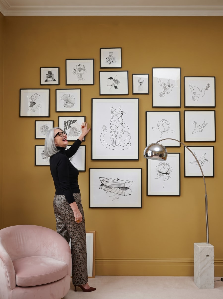 Her own one-line cat, framed at the centre of a full gallery wall — she is still laughing at it, hand half-raised from hanging it