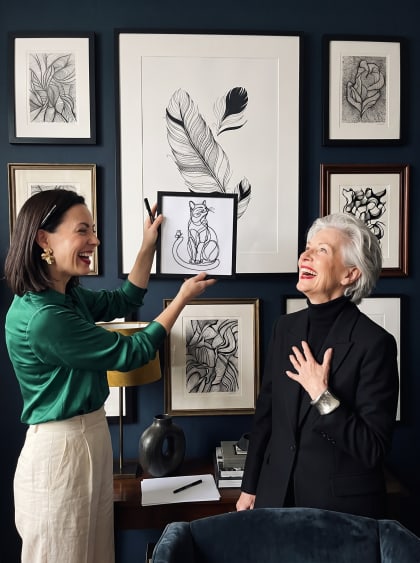 A deep navy gallery wall of framed ink drawings — one woman holds up her freshly drawn cat, pen still in hand, as her friend laughs