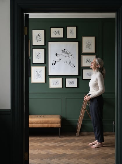 A forest-green panelled room — she laughs, picture wire still in hand, as her hare sits level