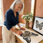 A woman filling a large butterfly zendoodle in black, ink blue and gold at a console, one already framed beside her