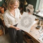 A finished botanical monogram wreath drawing held up in a plaster-walled studio