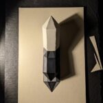Crystal drawing cover: a hand-cut paper solid under one lamp, each face a different flat tone, standing on its own ink drawing