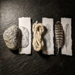 A stone, a coil of wool and a feather laid on dark slate, each touching a torn paper strip showing that same surface drawn in ink