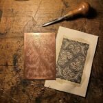 A copper plate cut with filigree scrollwork lying beside the damp sheet just printed from it, the same scrolls reversed on the metal