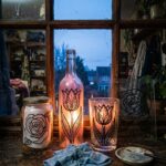 Glass painting ideas for beginners: painted jars, a bottle and a flat pane glowing on a windowsill at dusk