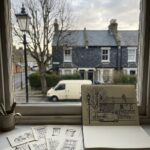 Urban sketching for beginners: a sketchbook on a kitchen windowsill showing the same street that is visible through the glass